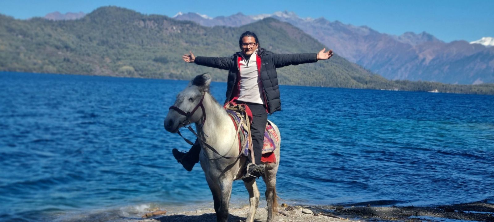 Hikmat on a white horse with arms outstretched at the shore of a deep-blue mountain lake