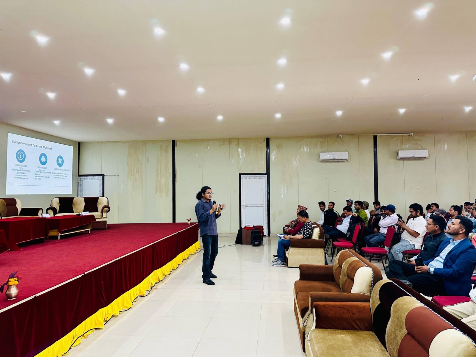 Hikmat speaking with a microphone to a seated audience beside a projected slide