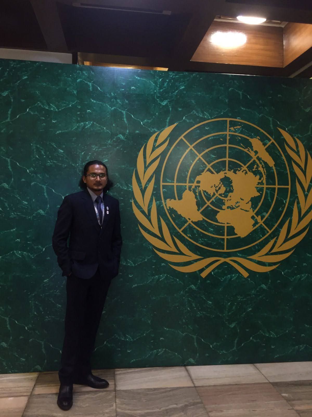Hikmat in a suit standing beside the United Nations emblem on a green marble wall